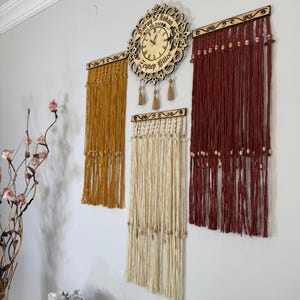 May include: A wall-mounted wooden clock with a decorative floral design, accompanied by three macrame wall hangings in mustard yellow, cream, and burgundy. The clock has a light-colored face and a wooden frame.