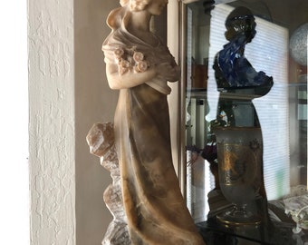 Puede incluir: Una escultura de alabastro blanco de una mujer sosteniendo flores, de pie sobre una base de mármol blanco. La escultura está en un estante con una puerta de vidrio.