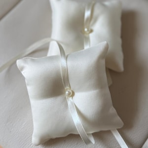 May include: Two ivory satin ring bearer pillows. Each square pillow features a pearl accent and a satin ribbon. The pillows are designed for a wedding ceremony.