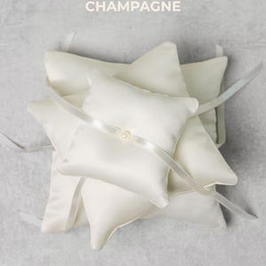 May include: Champagne-colored ring pillows. The pillows are square with pointed corners and are made of a satin-like material. A white ribbon is tied around each pillow, and a small pearl is attached to the ribbon. The word "CHAMPAGNE" is printed at the top.