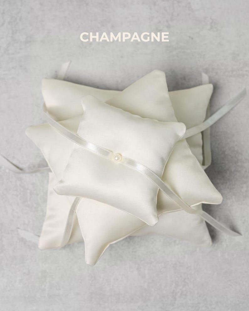 May include: Champagne-colored ring pillows. The pillows are square with pointed corners and are made of a satin-like material. A white ribbon is tied around each pillow, and a small pearl is attached to the ribbon. The word "CHAMPAGNE" is printed at the top.