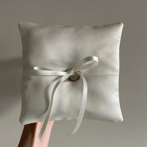May include: A white satin ring pillow with a white satin ribbon tied in a bow with a silver ring holder in the center.