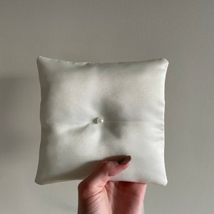 May include: Ivory satin ring pillow with a single pearl accent. The square pillow is held up against a neutral background. The pillow is a wedding accessory.
