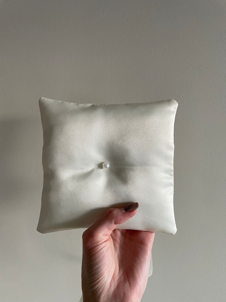 May include: Ivory satin ring pillow with a single pearl accent. The square pillow is held up against a neutral background. The pillow is a wedding accessory.