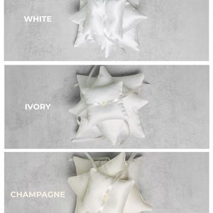 May include: Three ring bearer pillows in white, ivory, and champagne satin. Each pillow is adorned with a ribbon and a pearl. The word "WHITE", "IVORY", and "CHAMPAGNE" are printed above each pillow.