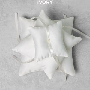 May include: Ivory-colored ring pillows with a star-like arrangement. Each pillow is square with a pointed edge and a satin ribbon. A pearl bead accents the center pillow. The word "IVORY" is printed at the top.