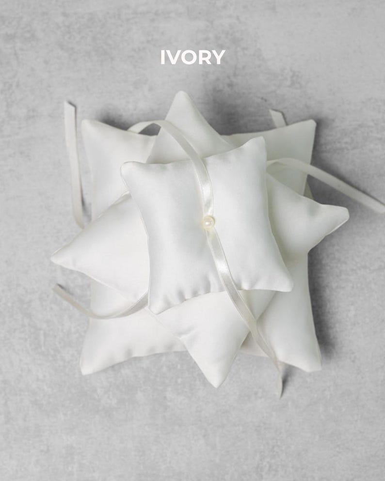 May include: Ivory-colored ring pillows with a star-like arrangement. Each pillow is square with a pointed edge and a satin ribbon. A pearl bead accents the center pillow. The word "IVORY" is printed at the top.