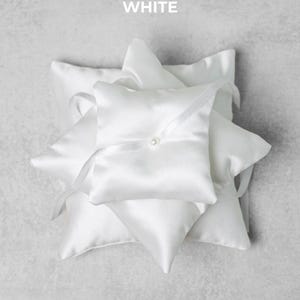 May include: A stack of white satin ring bearer pillows. Each pillow is square with a pointed corner and a ribbon tie. A small pearl is sewn onto the center of the top pillow. The word "WHITE" is printed at the top.