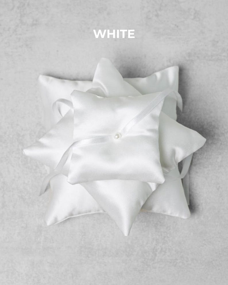 May include: A stack of white satin ring bearer pillows. Each pillow is square with a pointed corner and a ribbon tie. A small pearl is sewn onto the center of the top pillow. The word "WHITE" is printed at the top.