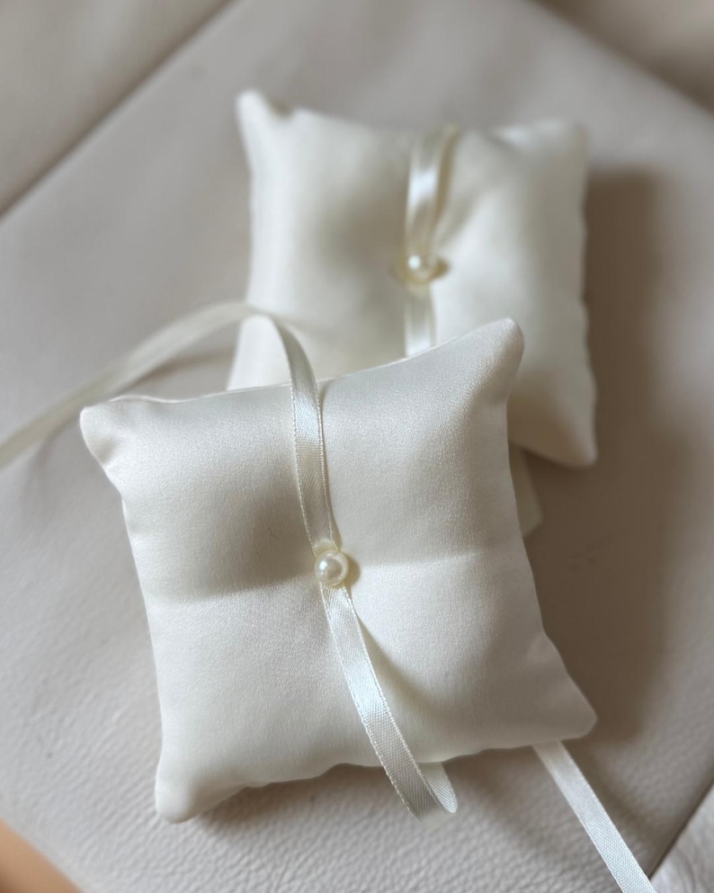 May include: Two ivory satin ring bearer pillows. Each square pillow features a pearl accent and a satin ribbon. The pillows are designed for a wedding ceremony.