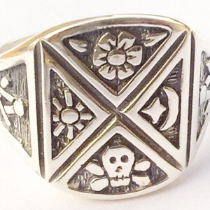 May include: A silver ring with a square design featuring four different symbols: a flower, a sun, a crescent moon with a star, and a skull and crossbones.