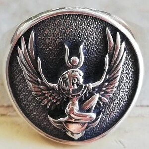May include: A silver ring with a detailed, raised design of a winged figure within a circular frame. The figure has outstretched wings and intricate detailing, set against a textured background. The ring has a polished, rounded band.