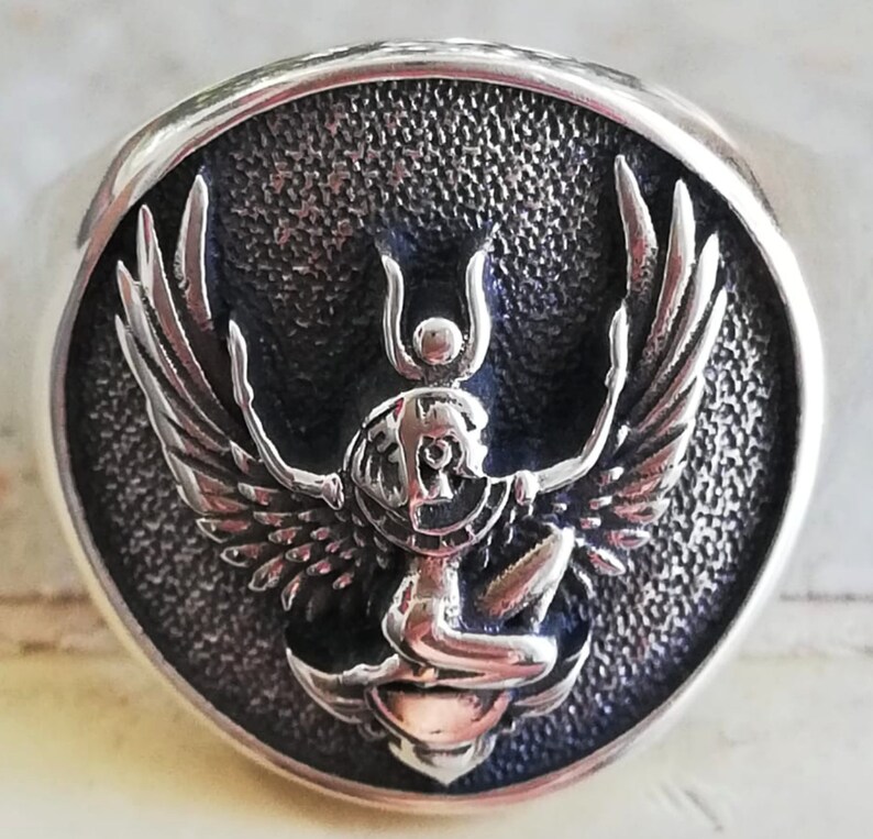 May include: A silver ring with a detailed, raised design of a winged figure within a circular frame. The figure has outstretched wings and intricate detailing, set against a textured background. The ring has a polished, rounded band.