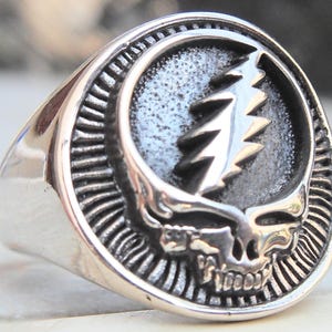May include: A silver-colored ring featuring a detailed skull design with a lightning bolt emblem. The ring has a round face with a textured background and a polished band. The design is reminiscent of a classic rock and roll aesthetic.