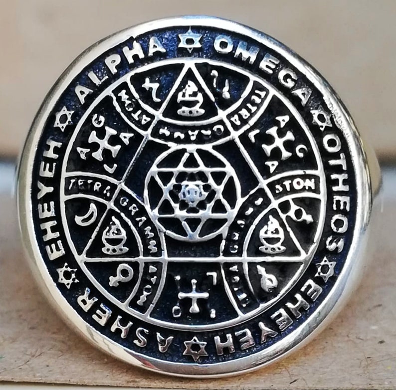 Puede incluir: Un anillo de plata con un dise&ntilde;o de esmalte negro que presenta una estrella de David, un tri&aacute;ngulo y las palabras "Alpha Omega Eheyieh Asheyr Etheos Otheos" alrededor del borde.