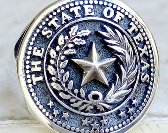 May include: A silver ring with a raised design of the State of Texas seal. The seal features a five-pointed star surrounded by a wreath of olive branches. The words "THE STATE OF TEXAS" are inscribed around the outer edge of the seal.