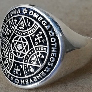 Puede incluir: Un anillo de plata con un dise&ntilde;o de esmalte negro que presenta una estrella de David, una cruz y otros s&iacute;mbolos. El anillo est&aacute; inscrito con las palabras "ALPHA & OMEGA COTHEC ASHER" y "GRAMMATON".