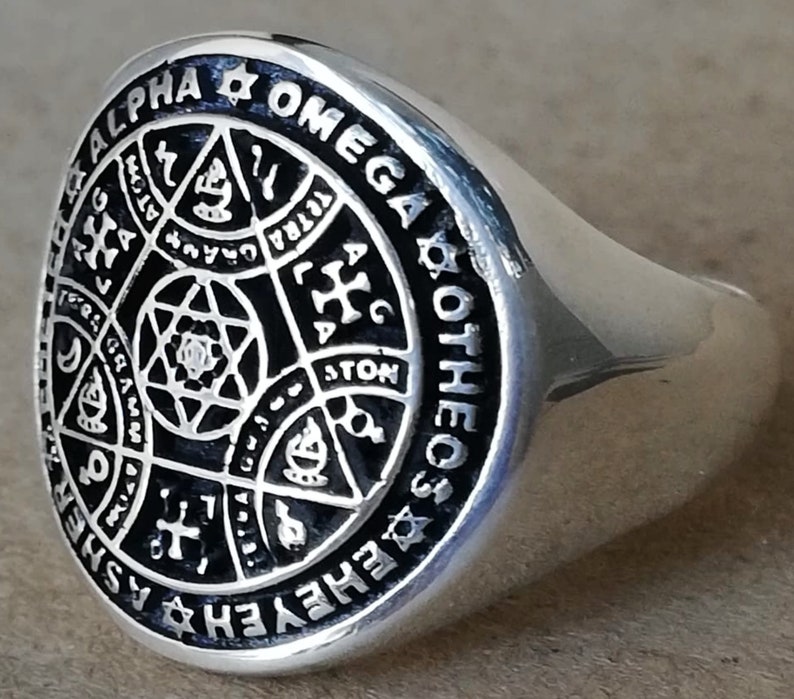 Puede incluir: Un anillo de plata con un dise&ntilde;o de esmalte negro que presenta una estrella de David, una cruz y otros s&iacute;mbolos. El anillo est&aacute; inscrito con las palabras "ALPHA & OMEGA COTHEC ASHER" y "GRAMMATON".