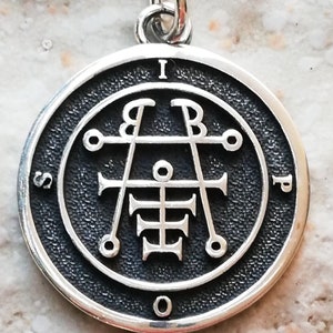 May include: A silver pendant with a black background and a silver sigil in the center. The sigil is surrounded by a circle with the letters "I.S.P.O." around it.