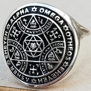 Puede incluir: Un anillo de plata con un dise&ntilde;o de esmalte negro que presenta una estrella de David, una cruz y otros s&iacute;mbolos. El anillo est&aacute; inscrito con las palabras "Alpha Omega Otheos" y "Ehedasher".