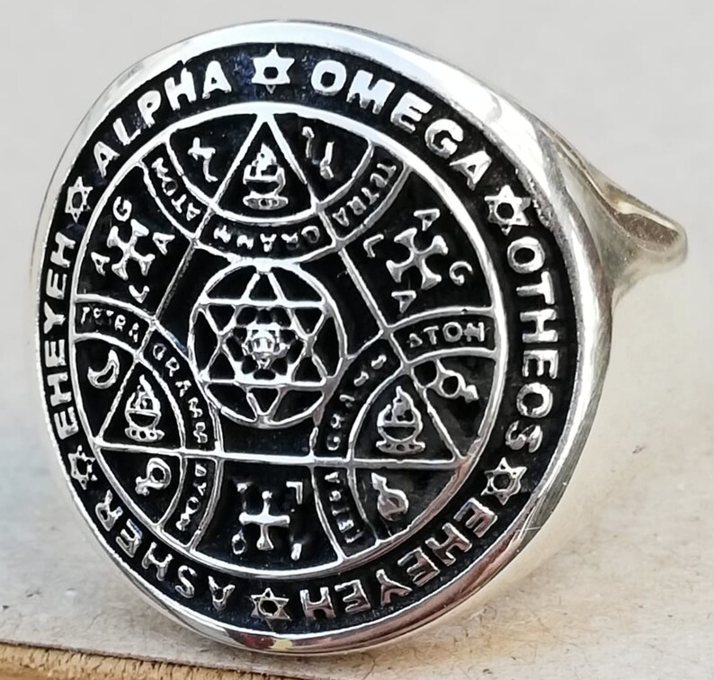 Puede incluir: Un anillo de plata con un dise&ntilde;o de esmalte negro que presenta una estrella de David, una cruz y otros s&iacute;mbolos. El anillo est&aacute; inscrito con las palabras "Alpha Omega Otheos" y "Ehedasher".