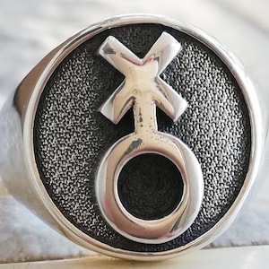 Könnte beinhalten: Ein silberner Ring mit einer strukturierten Oberfläche und einem erhabenen Symbol eines Kreises mit einem Kreuz darauf. Das Symbol befindet sich in der Mitte des Rings.
