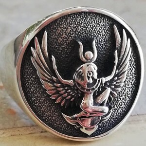May include: A silver ring featuring a detailed, raised design of a winged figure with a headdress, set against a textured, dark background. The ring has a wide band and a polished surface, highlighting the intricate details of the central emblem.