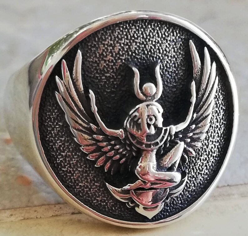 May include: A silver ring featuring a detailed, raised design of a winged figure with a headdress, set against a textured, dark background. The ring has a wide band and a polished surface, highlighting the intricate details of the central emblem.