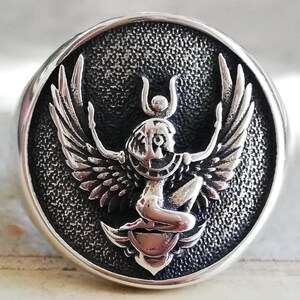 May include: A silver ring with a detailed, raised design of a winged figure with a headdress, set within a textured, circular frame. The ring has a polished, rounded band.