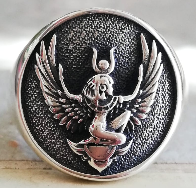 May include: A silver ring with a detailed, raised design of a winged figure with a headdress, set within a textured, circular frame. The ring has a polished, rounded band.