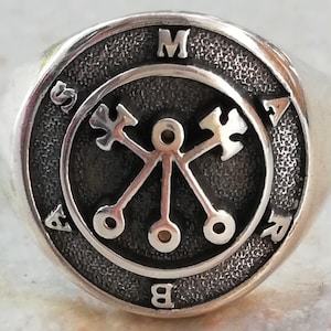 May include: A silver ring with a black circular design featuring a symbol with four arms and three circles. The letters "MASAB" are inscribed around the outer edge of the design.