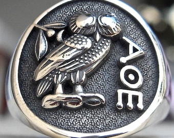 May include: A silver ring featuring a detailed owl design with the Greek letters AOE. The owl is perched on a branch with leaves, set against a textured background. The ring has a polished, rounded edge.