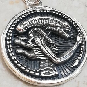 May include: Silver pendant featuring a detailed, three-dimensional alien creature design. The alien is depicted in a crouched position with its tail curled around its body. The pendant is circular and has a textured border.