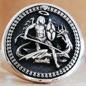 May include: A silver ring with a detailed design of an angel fighting a dragon. The angel is holding a sword and shield, and the dragon is wrapped around the angel's legs. The ring is surrounded by a chain-link border.
