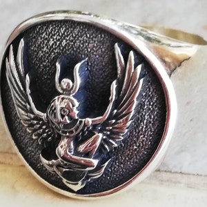 May include: A silver ring with a detailed relief of a winged figure with outstretched arms. The figure is set against a textured, dark background within an oval frame. The ring band is smooth and polished.