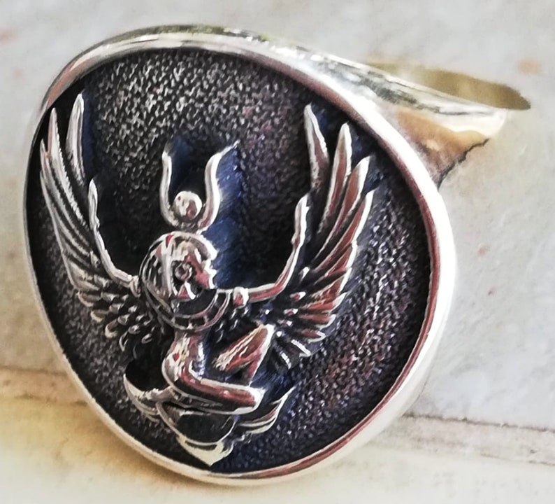 May include: A silver ring with a detailed relief of a winged figure with outstretched arms. The figure is set against a textured, dark background within an oval frame. The ring band is smooth and polished.