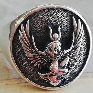 May include: A silver ring featuring a detailed, raised design of a winged figure with a headdress. The figure is set against a textured, dark background within a circular frame. The ring has a polished, shiny finish.