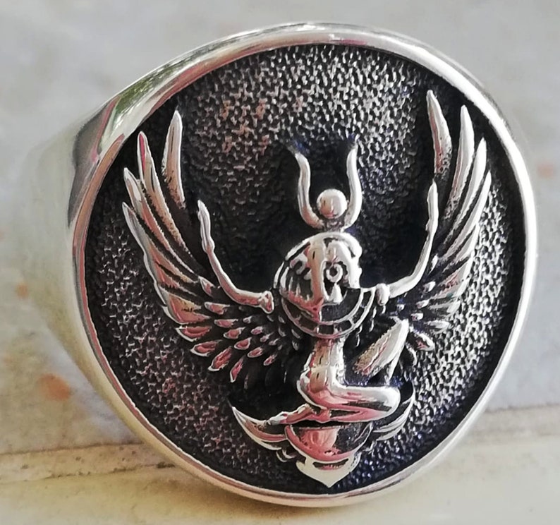 May include: A silver ring featuring a detailed, raised design of a winged figure with a headdress. The figure is set against a textured, dark background within a circular frame. The ring has a polished, shiny finish.
