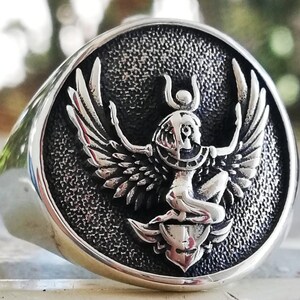 May include: A silver ring featuring a detailed relief of a winged figure, possibly an Egyptian deity. The ring has a polished, rounded band and a textured, circular face. The figure has outstretched wings and is seated above a stylized emblem.