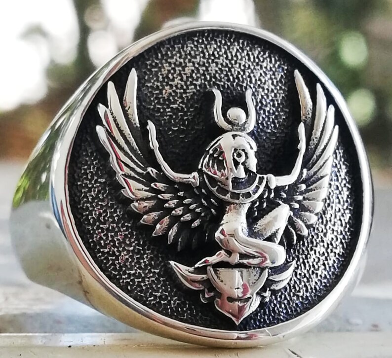 May include: A silver ring featuring a detailed relief of a winged figure, possibly an Egyptian deity. The ring has a polished, rounded band and a textured, circular face. The figure has outstretched wings and is seated above a stylized emblem.