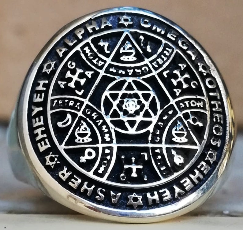 Puede incluir: Un anillo de plata con un dise&ntilde;o de esmalte negro que presenta un c&iacute;rculo con las palabras "ALPHA", "OMEGA", "COTHEOS", "ASHEREHEYEH" y "TETRA GRAMM" alrededor del borde. El centro del c&iacute;rculo presenta una estrella de David con un c&iacute;rculo m&aacute;s peque&ntilde;o en su interior.