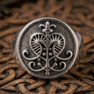 Könnte beinhalten: Ein silberner Ring mit einem detaillierten, symmetrischen Design. Der Ring zeigt ein zentrales Emblem mit geschwungenen Elementen und einer kronenartigen Spitze. Der Ring ist vor einem Holzgrund platziert, was seine komplizierten Details hervorhebt.