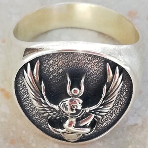 May include: A silver ring with an oval face depicting a detailed relief of a winged deity. The deity has a human-like head and outstretched wings. The ring has a polished finish.