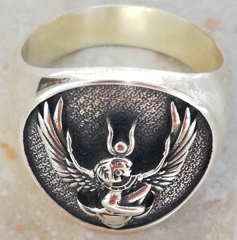 May include: A silver ring with an oval face depicting a detailed relief of a winged deity. The deity has a human-like head and outstretched wings. The ring has a polished finish.