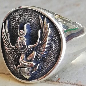 May include: A silver signet ring with an oval face. The face features a detailed relief of a winged figure with a headdress, set against a textured, dark blue background. The ring has a polished, reflective finish.