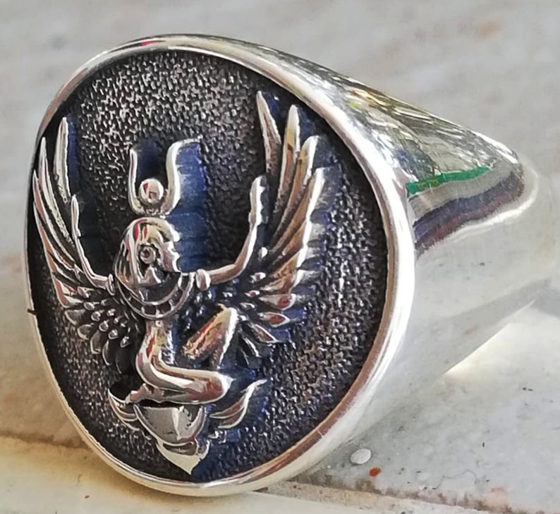 May include: A silver signet ring with an oval face. The face features a detailed relief of a winged figure with a headdress, set against a textured, dark blue background. The ring has a polished, reflective finish.