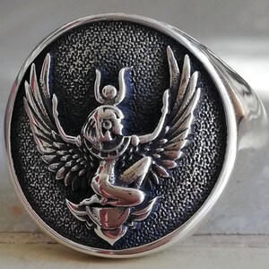 May include: A silver ring featuring a detailed relief of a winged figure, possibly an Egyptian deity, set against a textured, dark backdrop. The ring has a polished, rounded band and a circular face. The figure has outstretched wings and a headdress.