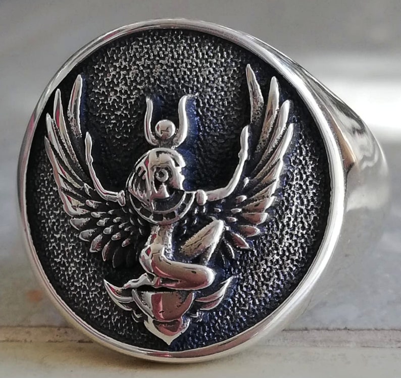 May include: A silver ring featuring a detailed relief of a winged figure, possibly an Egyptian deity, set against a textured, dark backdrop. The ring has a polished, rounded band and a circular face. The figure has outstretched wings and a headdress.