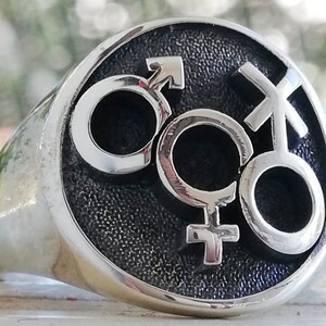 Puede incluir: Un anillo plateado con una cara redonda que presenta tres símbolos de género: masculino, femenino e intersexual. Los símbolos están en relieve sobre un fondo oscuro y texturizado. El anillo tiene una banda pulida y redondeada.
