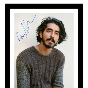May include: A framed photograph of a man with dark hair and a mustache, wearing a textured grey jumper and blue trousers. The image is signed in blue ink. The photograph is set against a neutral background.